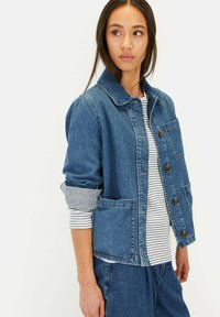 Jeansjacke mit Kragen, Knopfleiste, zwei Fronttaschen und aufgerollten Ärmeln. Die Farbe ist Mittelblau mit einer strukturierten Oberfläche.