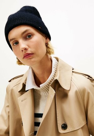 Beige trench coat with black buttons, layered over a cream sweater with navy stripes. Model wears a navy beanie and a white collared shirt.