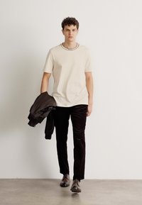T-shirt beige à manches courtes avec un col noir contrastant, associé à un pantalon en velours noir et une veste noire drapée sur le bras, chaussures brunes.