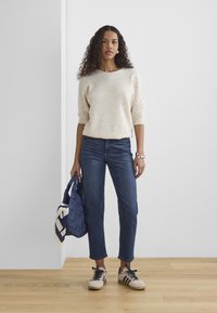 Pull crème avec des manches bouffantes texturées, associé à un jean bleu foncé. Tient un sac en toile bleu et porte des baskets beiges avec des accents noirs.