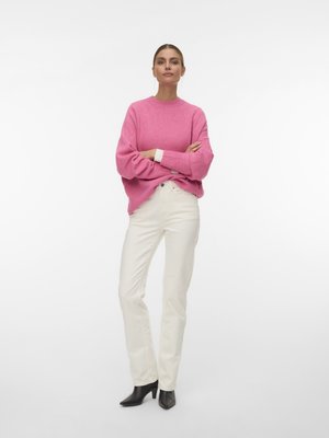 Γυναίκα στέκεται με σταυρωμένα τα χέρια φορώντας ροζ πουλόβερ, λευκό παντελόνι με ψηλή μέση, μαύρα μποτάκια στον αστράγαλο και κρεμαστά σκουλαρίκια σε λευκό φόντο.