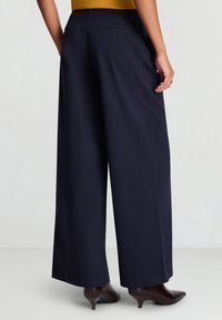 Weit geschnittene Hosen aus marineblauem Stoff mit klarem Design, Schritt-Details und Gesäßtaschen, kombiniert mit dunklen Absatzschuhen.