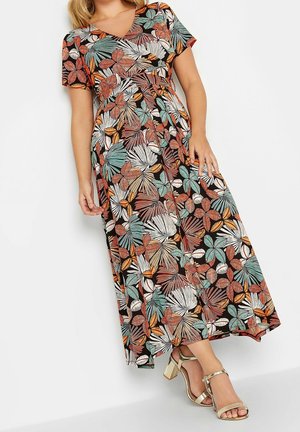 Vrouw die een kleurrijke maxi-jurk met korte mouwen en een bladmotief draagt, met een V-hals en gouden sandalen met hoge hakken, staand tegen een witte achtergrond.