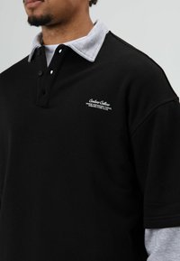 Zwart poloshirt met een grijze, getextureerde kraag, voorzien van een drieknopsplooi en een geborduurd wit logo op de linkerkant van de borst. Zachte stof.