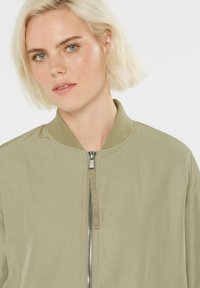 Olivgrüne Bomberjacke mit geripptem Kragen und silbernem Reißverschluss. Glatter Stoff mit minimalen Designdetails. Nahaufnahme des Gesichts des Models.