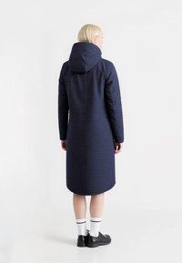 Lang marineblauwe jas met een capuchon, gemaakt van gestructureerde stof. Heeft een rechte snit en valt onder de knie. Gekleed met zwarte schoenen en witte sokken.