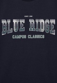 Jūras zila kokvilnas T-kreklā ir izcili balti un zaļi uzraksti: "BLUE RIDGE" ar šķīru rakstu, "CAMPUS CLASSICS" un "SINCE 1986."