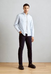 Camisa de rayas azul claro con botones, camiseta blanca debajo, jeans oscuros, zapatos negros. Atuendo casual con un ajuste relajado y detalles mínimos.