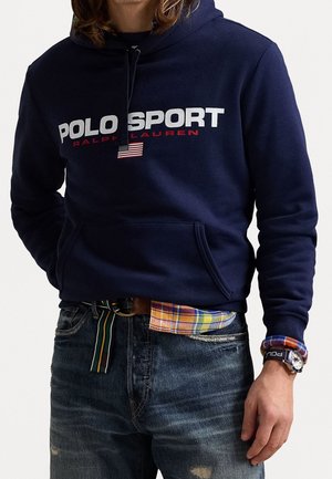 Person trägt eine marineblaue Polo Sport Kapuzenjacke mit amerikanischer Flagge, Hemdmanschette mit Karomuster, gestreiften Gürtel, abgenutzte Jeans und Armbanduhr.