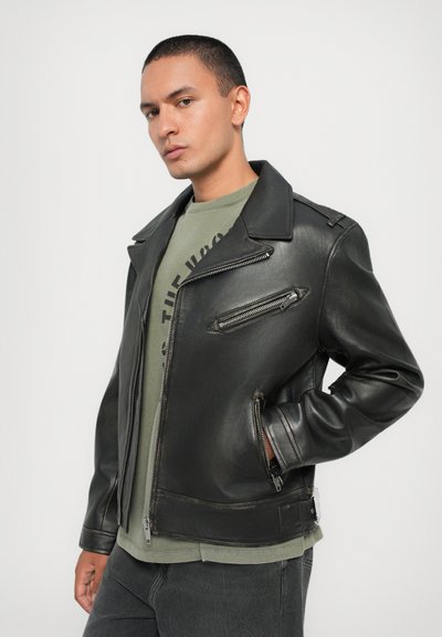 Μαύρο δερμάτινο μπουφάν μοτοσικλέτας με ευρύ γιακά, πολλαπλές ζιπ-πορτ, και λεία υφή, στρωμένο πάνω από πράσινη μπλούζα.