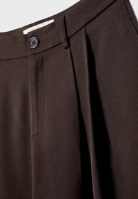 Gros plan d’un pantalon marron foncé mettant en valeur la ceinture avec bouton, passants et pli avant.