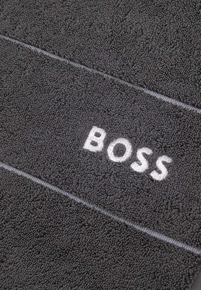 Mörkgrå handduk med texturerat, fluffigt tyg. Har en vit broderad "BOSS"-logotyp och ett subtilt linjemönster över ytan.
