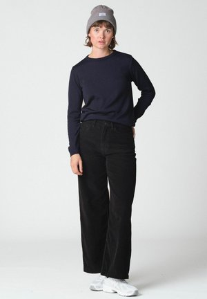 Jeune personne portant un bonnet gris, un t-shirt à manches longues sombre, un pantalon noir à jambes larges et des baskets blanches, debout devant un fond uni.
