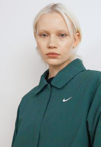 Zöld Nike dzseki sima textúrával, hegyes gallérral és fehér Nike logóval a bal mellen. Egyszerű design, könnyű anyag.