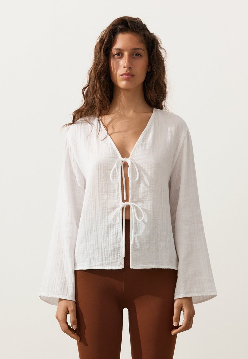 Femme aux longs cheveux bouclés portant un chemisier blanc à manches longues noué à l'avant et un pantalon marron ajusté, debout devant un fond uni.