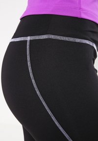 Czarne legginsy sportowe o gładkiej fakturze, z kontrastowym białym szwem i częściowym widokiem fioletowej bluzki.