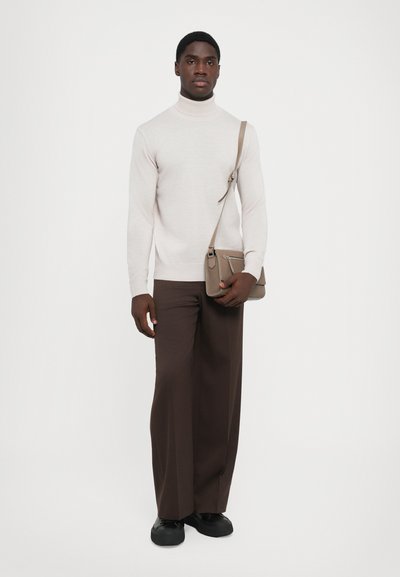Maglione a collo alto color beige chiaro abbinato a pantaloni a gamba larga marrone scuro e scarpe nere; la modella tiene in mano una piccola borsa a tracolla marrone.