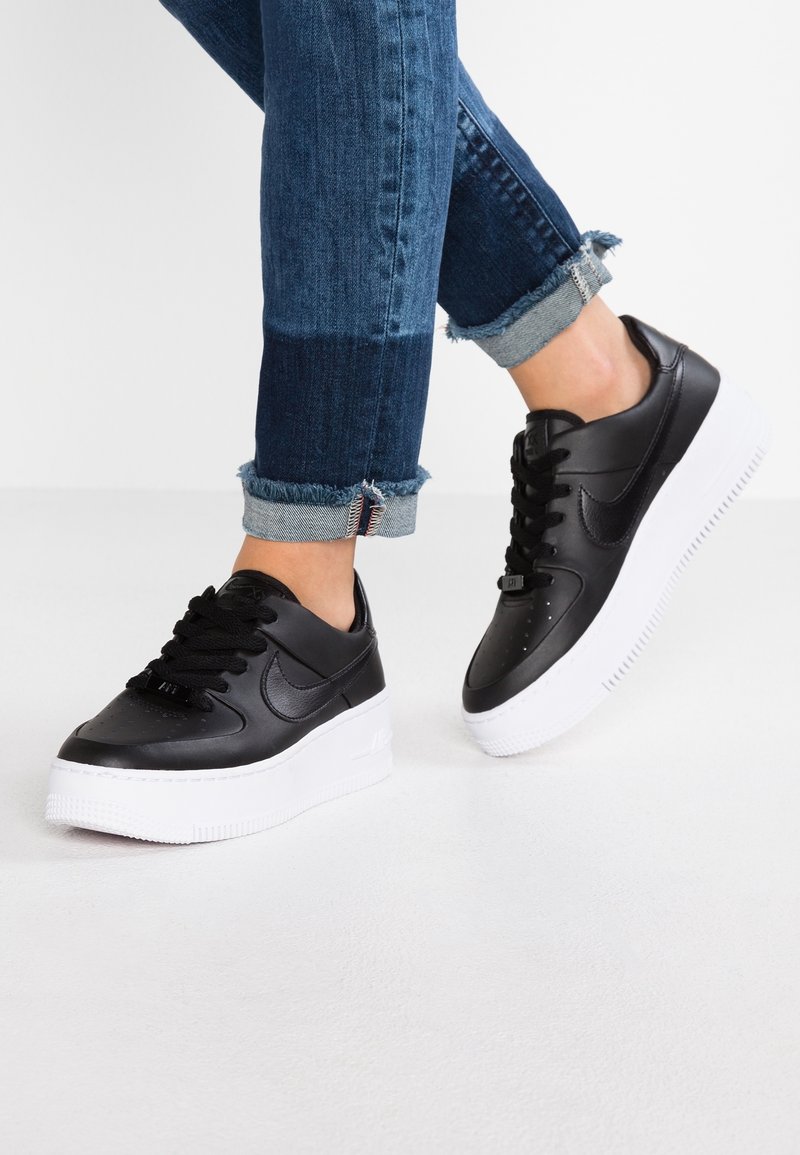 Personne portant un jean bleu foncé retroussé et des baskets en cuir noir avec des semelles blanches épaisses, debout sur un sol blanc.
