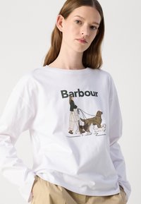 T-shirt blanc à manches longues avec un imprimé graphique d'une personne promenant des chiens. Le design comprend des couleurs atténuées et une coupe décontractée.