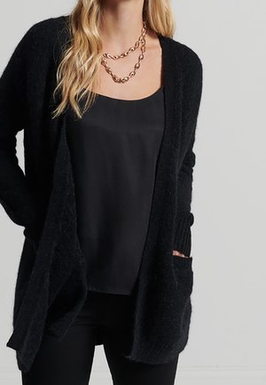 Femme portant un cardigan noir duveteux ouvert sur un caraco en satin noir, accessoirisé d’un collier chaîne épaisse dorée.