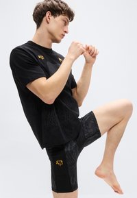 Camiseta negra con detalles dorados y un patrón sutil combinada con pantalones cortos de compresión negros que presentan un diseño texturizado y un emblema dorado.