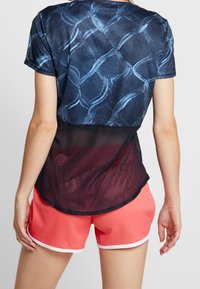 Kobieta ubrana w niebieską bluzkę z krótkim rękawem w wzory z czarną siateczkową tylną częścią oraz koralowe spodenki sportowe z białym wykończeniem, stojąca tyłem.