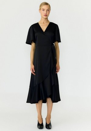 Mulher com cabelo louro vestindo um vestido preto com mangas curtas e fluídas e saltos pretos, em pé contra um fundo branco simples.