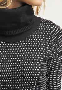 Pull à col roulé noir avec un motif texturé de petits triangles blancs. Matière douce et tricotée avec des détails côtelés au niveau du col.