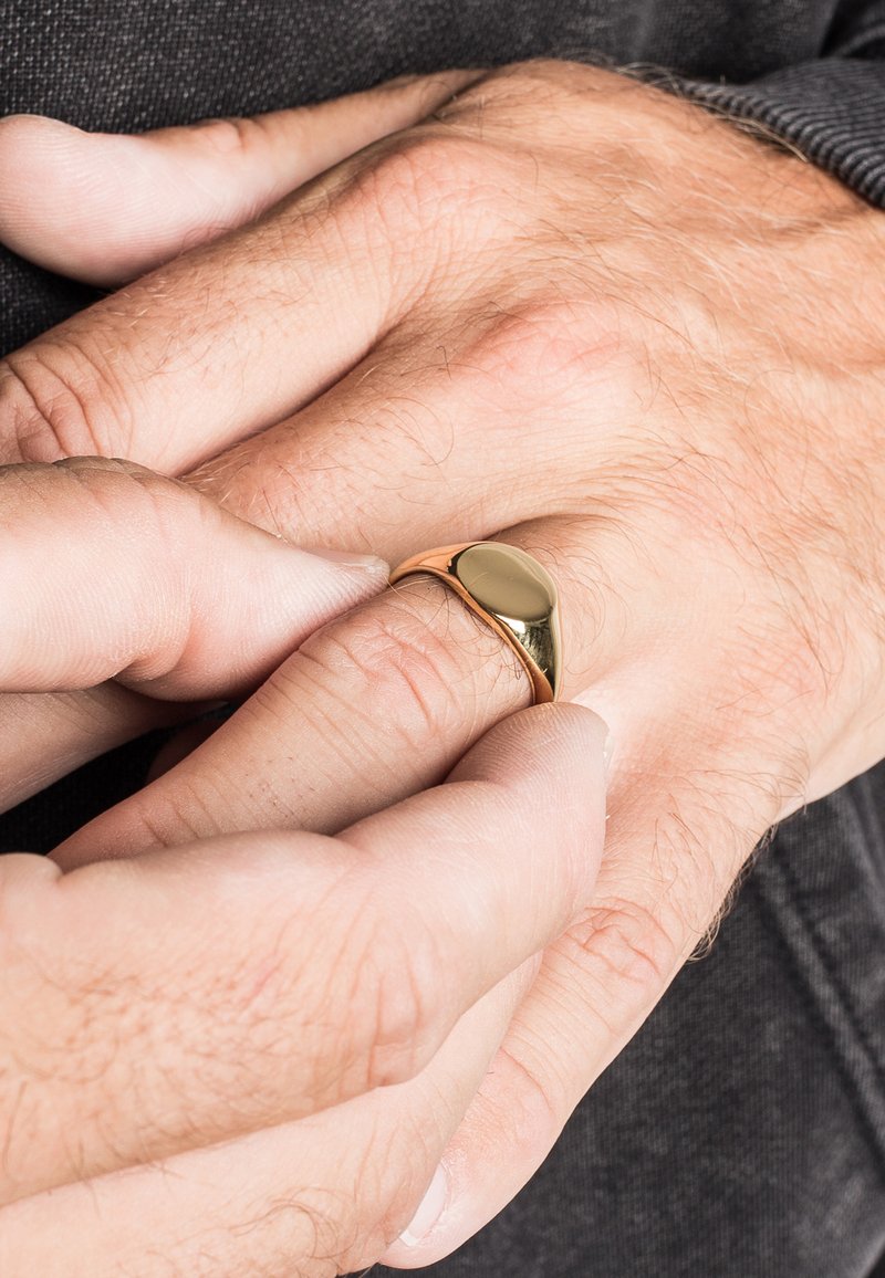 Gouden ring met een glad, afgerond ontwerp zit op een vinger, met handen in de nabijheid, waarbij de huidtextuur en subtiele details worden getoond.