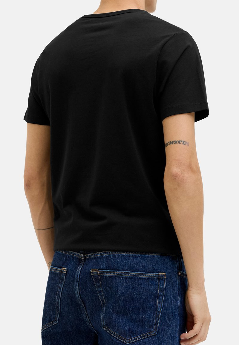 Personne portant un t-shirt noir uni à manches courtes et un jean en denim bleu, vue de dos avec un tatouage de chiffres romains visible sur le bras droit.