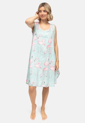 Vrouw draagt een mouwloze lichtblauwe jurk met roze flamingoprint, staat blootsvoets en raakt met één hand haar haar aan.