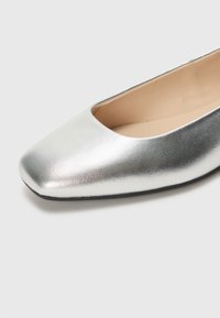 Zapato plano plateado con puntera cuadrada, hecho de cuero metálico, con una textura suave y diseño minimalista. La suela es negra y duradera.