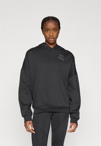 Crna majica s kapuljačom napravljena od mekanog materijala, s kapuljačom na vezanje, ležernim krojem i suptilnim logotipom na prsima. Uparena s crnim tajicama.