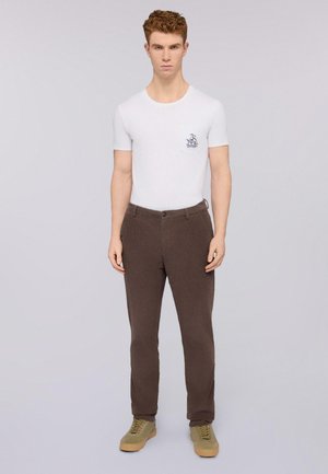 T-shirt blanche ajustée avec un détail de poche et un motif graphique, associée à un pantalon marron foncé. Le mannequin porte des baskets vert olive. Fond uni.