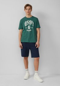 Zeleni pamučni T-shirt s bijelim tiskanim grafičkim elementima, u kombinaciji s tamnoplavim kratkim hlačama. Model nosi bijele tenisice s prugastim čarapama. Jednostavan dizajn.