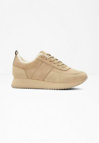 Baskets de sport beiges avec une tige en simili cuir lisse, une semelle texturée, un laçage à l'avant et une doublure douillette visible au niveau de la cheville.