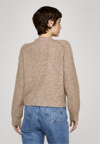 Brun, teksturert genser med kort design og ribbestrikkede mansjetter, kombinert med høytlivs blå jeans og ensfarget bakgrunn.