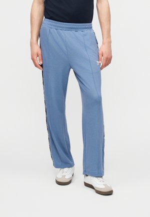 Άνδρας με ανοιχτό μπλε φόρμα με μαύρες και λευκές πλαϊνές ρίγες και λευκά αθλητικά παπούτσια, στέκεται μπροστά σε απλό φόντο.