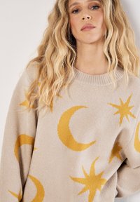 Beige lange mouwtrui met gele halve maan- en sterrenpatronen. Zachte textuur met een ronde halslijn en verlaagde schouders.