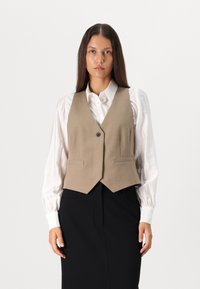 Beige uldblandingsvest med V-hals, enkelt knaplukning og to forlommer, båret over en hvid bluse og sort nederdel.