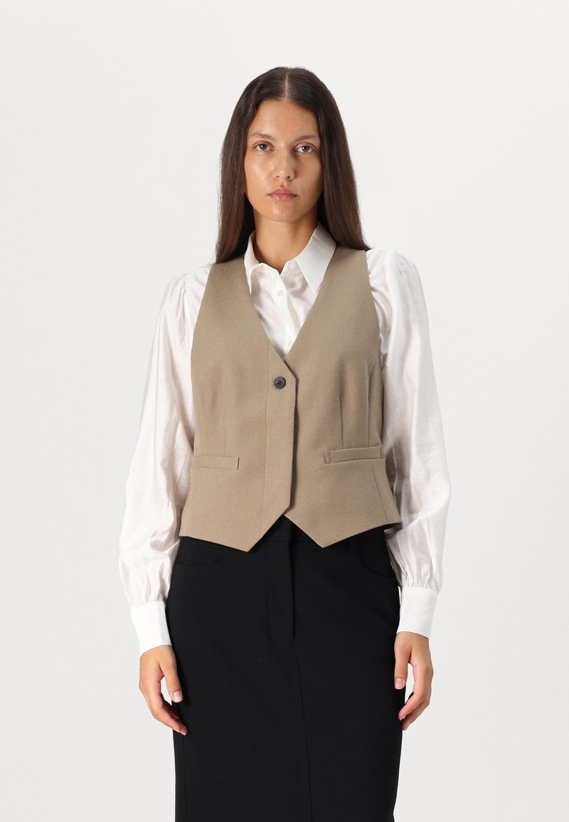 Beige uldblandingsvest med V-hals, enkelt knaplukning og to forlommer, båret over en hvid bluse og sort nederdel.