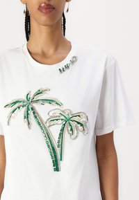 T-shirt bianco in cotone con ricamo di una palma verde e dettagli in gioiello. Collo rotondo con decorazioni. Maniche corte.