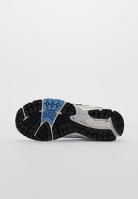 Svart gummi yttersula med texturerat grepp, blå detaljer och ett centralt mönster. Har en vit mellansula och ljus ovandel.