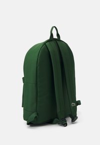 Sac à dos en tissu vert avec une forme arrondie, des bretelles rembourrées, une poche avant et un détail de logo sur le côté. Texture lisse.