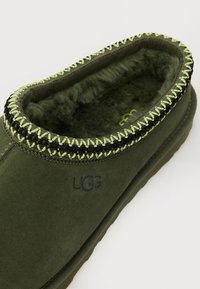 Chausson en daim vert avec une couture tressée noire et verte sur le bord, muni d'une doublure intérieure douce et d'un logo embossé sur le côté.