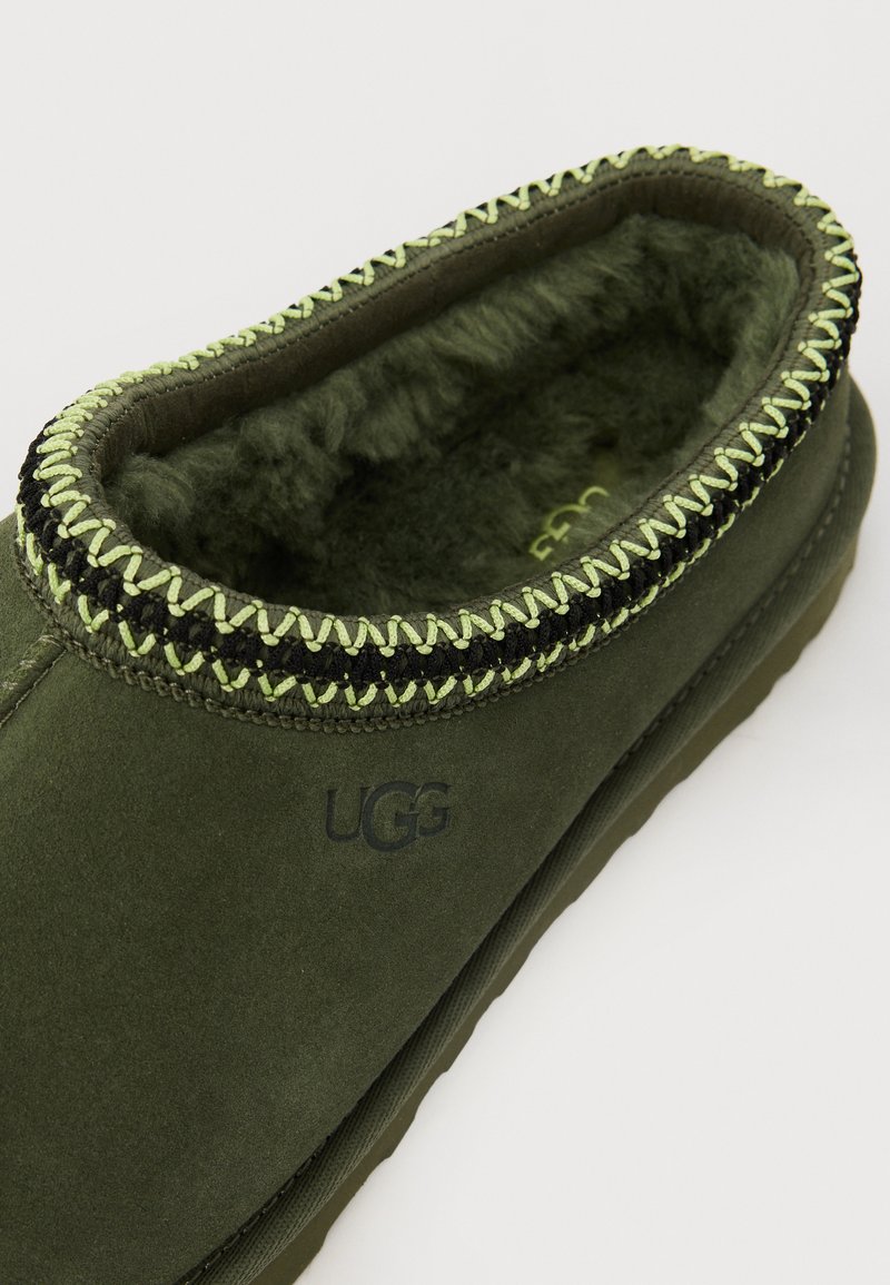 Groene suède slipper met zwart en groen gevlochten stiksel aan de rand, voorzien van een zachte binnenvoering en een verdiept logo aan de zijkant.