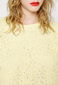 Haut jaune clair en dentelle avec motif floral, comportant des dessins complexes et une texture en maille ajourée, présentant un col rond.