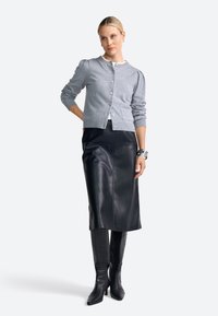 Cardigan en tricot gris clair avec un col rond, des boutons et des manches bouffantes, associé à une jupe midi en cuir noire et des bottes noires haute genou.