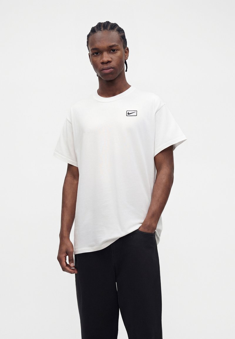 Junger Mann mit geflochtenem Haar, der ein weißes Nike-T-Shirt und schwarze Hose trägt, steht mit einer Hand in der Tasche vor einem einfarbigen Hintergrund.