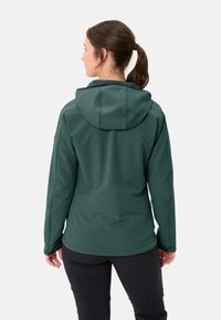 Giacca softshell verde scuro con cappuccio, caratterizzata da una texture liscia e un design snodato, indossata sopra pantaloni neri, vista da dietro.